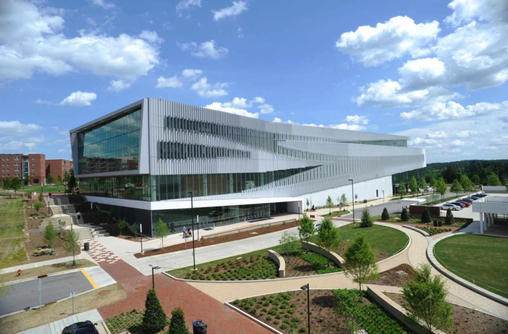 Centennial Campus main building with distinct modern architecture on the outside wall (made of glass and metal). It is surrounded by greenery, sidewalks, and a main parking lot.