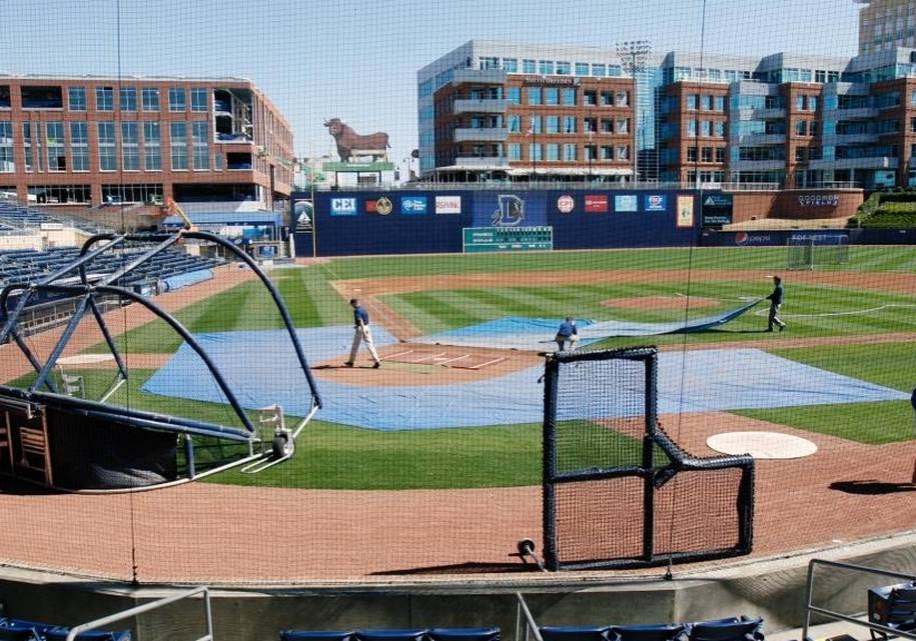 Durham Bulls stadium on a sunny afternoon. Crew members are removing rain tarps from the field.