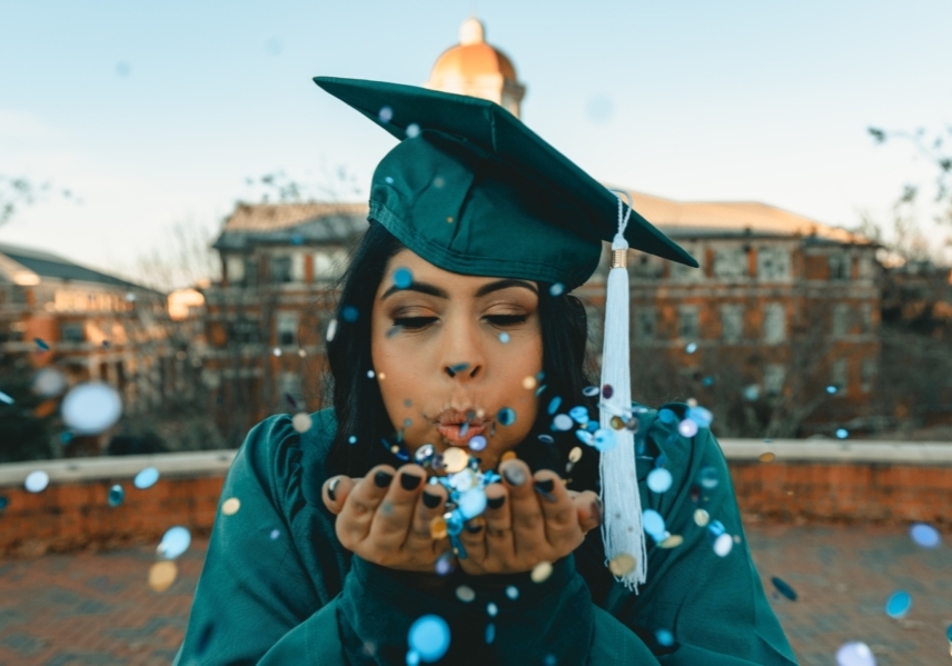 A young graduate in cap in gown. She is blowing confetti towards the camera from her outstretched hands.